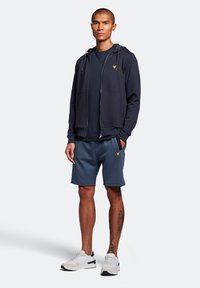 Felpa con zip di colore navy, dotata di coulisse, abbinata a una t-shirt blu scuro e pantaloni corti coordinati. Le sneakers presentano dettagli bianchi e neri, con un tatuaggio visibile.