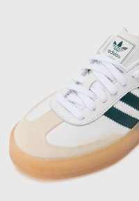 White leather sneakers with green stripes, suede toe cap, and gum sole. Features white laces and an Adidas logo on the tongue.