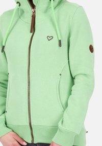 Sudadera con cremallera de color verde claro con capucha, que presenta un logo de corazón, puños elásticos y un detalle de cuero marrón en la manga. Tejido texturizado.