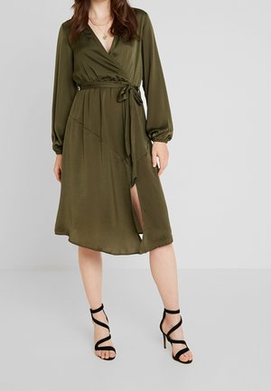 Kvinde iført en olivengrøn omslagskjole med lange ærmer og bindebånd i taljen, kombineret med sorte høje sandaler med remme.