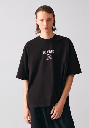 Jeune personne aux cheveux mi-longs porte un t-shirt noir oversize avec un texte rose et de petits cœurs sur la poitrine.