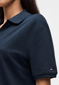 Γυναίκα που φοράει μπλε σκούρο κοντομάνικο πόλο με μικρό κόκκινο, άσπρο και μπλε λογότυπο σημαίας στο μανίκι.