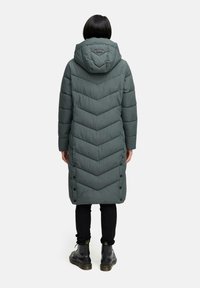 Cappotto imbottito verde lungo con cappuccio, cuciture a zigzag, bottoni a pressione lungo il lato e finitura opaca. Indossato sopra pantaloni neri e stivali.
