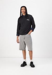 Felpa nera con una camicia di jeans blu a collo sotto, abbinata a pantaloni corti di jeans grigi chiari e scarpe nere con suola spessa.