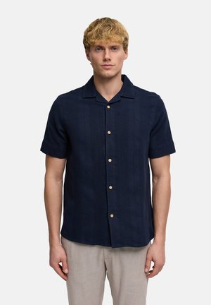 Blond man wearing a navy short-sleeve button-up shirt with light beige pants, standing facing forward on a plain background.