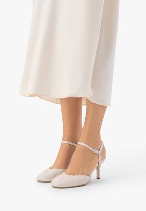 Stopy kobiety w beżowych czółenkach na obcasie z paskiem wokół kostki, w połączeniu z kremową, zwiewną sukienką.