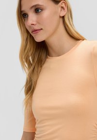 Hellpeachfarbener, taillierter T-Shirt mit kurzen Ärmeln, aus glattem Stoff gefertigt. Verfügt über einen Rundhalsausschnitt und dezente Rüschen an der Seite.