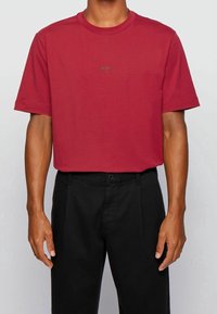 Červené tričko s kulatým výstřihem vyrobené z bavlny, s krátkými rukávy a nenápadným černým logem na přední straně. Nošeno s černými kalhotami.