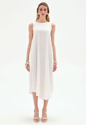 Femme portant une robe midi blanche asymétrique sans manches avec des bracelets dorés et des sandales dorées à talons hauts et brides, sur fond blanc.