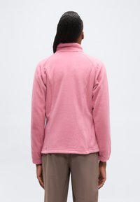 Άτομο που φοράει ροζ φλις μπουφάν με μακριά μανίκια και μπεζ παντελόνια, όρθιο με την πλάτη στραμμένη προς ένα απλό φωτεινό φόντο.