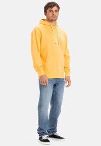Yellow hoodie with a kangaroo pocket, drawstrings, long sleeves, paired with light blue jeans and black sneakers on a plain background.