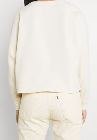 Persona che indossa un semplice top bianco sporco a maniche lunghe e pantaloni a righe beige chiaro, mostrata di spalle su uno sfondo bianco.