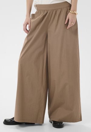 Luźne, szerokie spodnie w kolorze jasnobrązowym, noszone razem z czarnymi butami, białym topem oraz złotą bransoletką i pierścionkiem na prawej ręce.