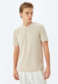 T-shirt beige a maniche corte realizzato in tessuto morbido, con scollo rotondo e taschino sul petto. Texture liscia con una vestibilità comoda.
