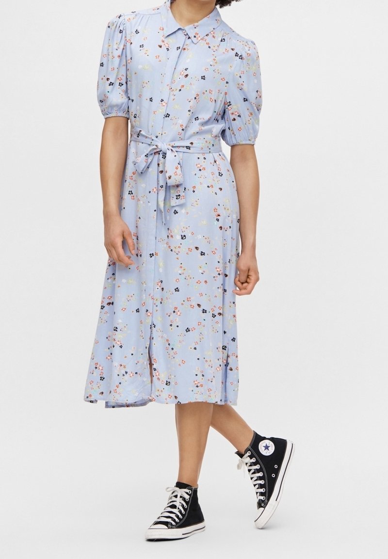 Robe boutonnée bleu clair à motifs floraux, avec manches courtes bouffantes, taille ceinturée et longueur midi, associée à des baskets montantes noires.