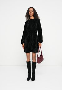 Musta samettimekko, jossa on pitkät hihat ja gathered-vyötärö. Yhdistetty polvipitkiin mustiin saappaisiin ja pieneen burgundin väriseen käsilaukkuun. Yksinkertainen tausta.