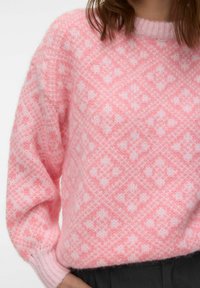 Femme portant un pull en tricot rose avec un motif de losanges et de fleurs, associé à un pantalon foncé, sur un fond blanc.