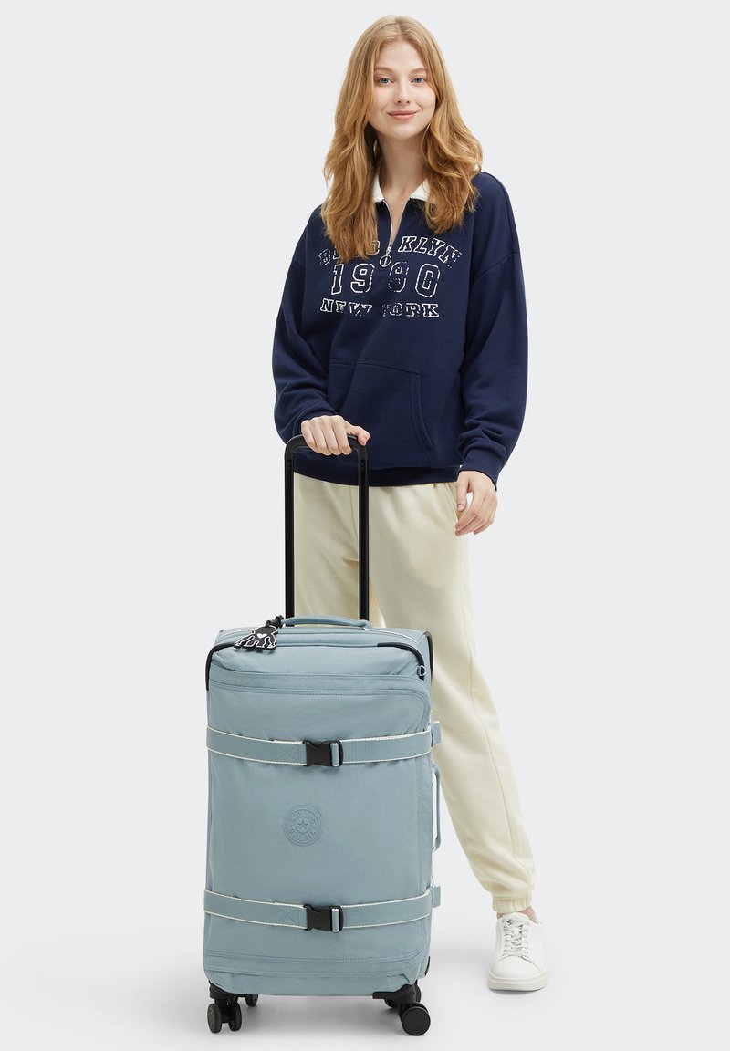 Maleta de viaje azul claro con dos ruedas negras, correas blancas y un logo. La maleta cuenta con un asa superior para facilitar el transporte.