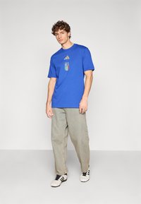 Tricou albastru din bumbac cu un logo Adidas auriu și un blazon al Italiei; modelul poartă blugi largi gri deschis și adidași alb-negru.