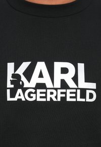 Camiseta negra con texto en blanco y en negrita "KARL LAGERFELD," que presenta una silueta de un perfil a la izquierda. Material de algodón con textura acanalada.