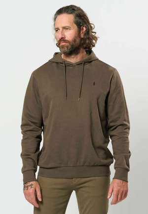 Homme avec barbe et cheveux longs portant un sweat à capuche vert olive et un pantalon assorti, debout devant un fond clair uni.