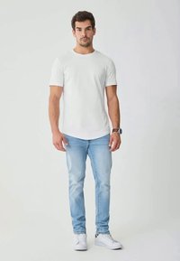 Mann som står iført hvit T-skjorte, lyseblå jeans, hvite joggesko og klokke mot en enkel lys bakgrunn.