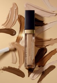 Corrector Futurist de Estée Lauder en un tubo transparente con tapa negra, rodeado de swatches de diversos tonos beige y marrón sobre un fondo dorado.