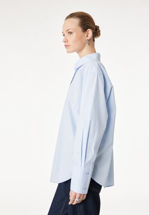 Vrouw met haar in een knot, draagt een lichtblauw oversized blouse met knopen, zilveren creolen en donkere broek, kijkt naar links tegen een witte achtergrond.