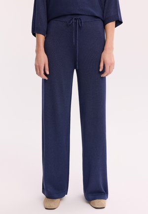 Granatowe, szerokie spodnie dzianinowe ze ściągaczem w talii, noszone z beżowymi, fakturowanymi butami o kwadratowym nosku na jasnym tle.