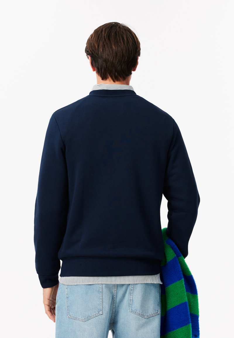 Granatowy sweter z prążkowanym dekoltem i mankietami, w połączeniu z koszulą w paski. W ręku trzymany jest zielono-niebieski szal w kratę.