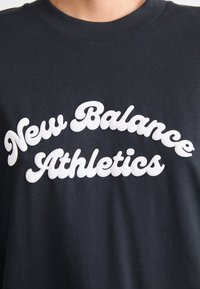 T-shirt noir à manches courtes avec un texte en caoutchouc blanc indiquant "New Balance Athletics" dans une police en gras et arrondie.