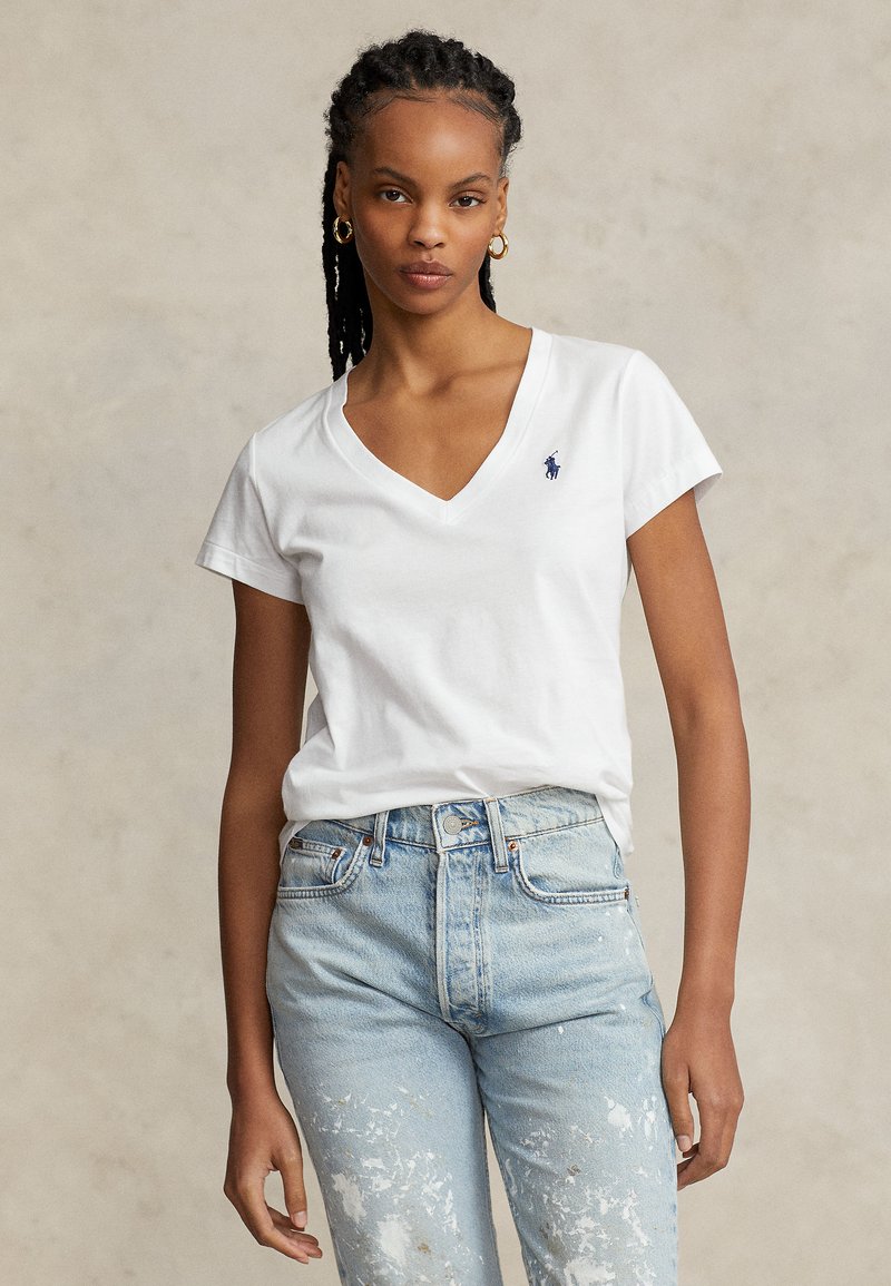 Kvinna med flätat hår som bär guldhoopörhängen, en vit V-ringad t-shirt med en liten blå logotyp och ljusblå jeans med vita färgsplatter.