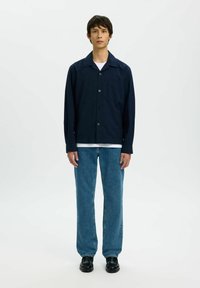 Chemise bleu marine boutonnée avec une poche, jean bleu clair ample, et chaussures noires. Le mannequin se tient devant un fond blanc uni.