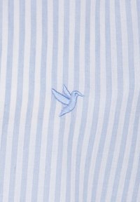 Tela a rayas azul claro con rayas blancas, que presenta un pequeño colibrí bordado en un azul más claro. Textura suave, diseño casual.