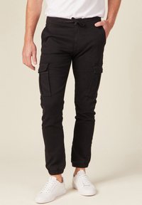 Zwarte cargobroek met verstelbare tailleband, voorzien van twee zijzakken en elastische boorden, gecombineerd met witte sneakers.