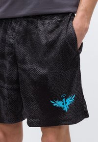 Μαύροι αθλητικοί σορτς με υφή, με μπλε λογότυπο σπαθιού με φτερά κοντά στο τελείωμα στο δεξί πόδι, φορεμένα με το δεξί χέρι στην τσέπη.