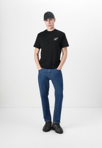 Svart bomull T-skjorte med logo, blå denimjeans med rett passform, og svarte joggesko med teksturert design. Modell står med hendene i lommene.