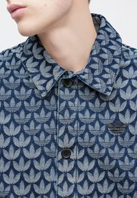 Jeans jakna z zapletenim vzorcem modrih in sivih rastlin, z gumbi za zapiranje in ikoničnim logotipom na prsih. Teksturni material.