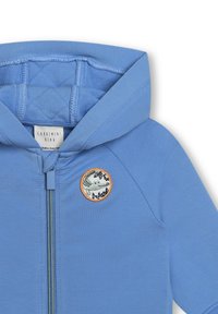 Felpa con zip blu, foderata con imbottitura trapuntata. Presenta un'etichetta ricamata rotonda che raffigura una rana, con accenti arancioni attorno all'etichetta.