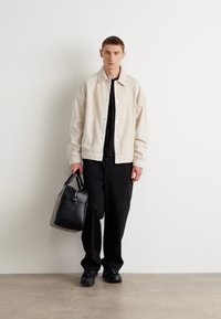Giacca bomber beige chiaro con colletto, t-shirt nera, pantaloni neri larghi e scarpe nere. In mano tiene una borsa a tracolla in pelle nera.