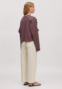 Langärmliges, locker sitzendes Oberteil in Mauve mit Rüschendetails, kombiniert mit hellbeigen weit geschnittenen Hosen und schwarzen Flip-Flops.