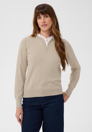 Femme aux longs cheveux bruns portant un pull beige par-dessus une chemise blanche à col en dentelle et un pantalon bleu foncé, debout devant un fond uni.