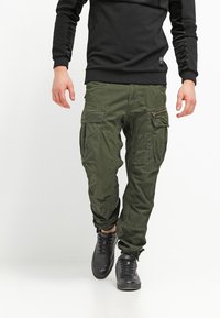 Gröna cargobyxor med flera fickor och dragkedjor, tillsammans med en svart långärmad tröja och svarta texturerade sneakers.
