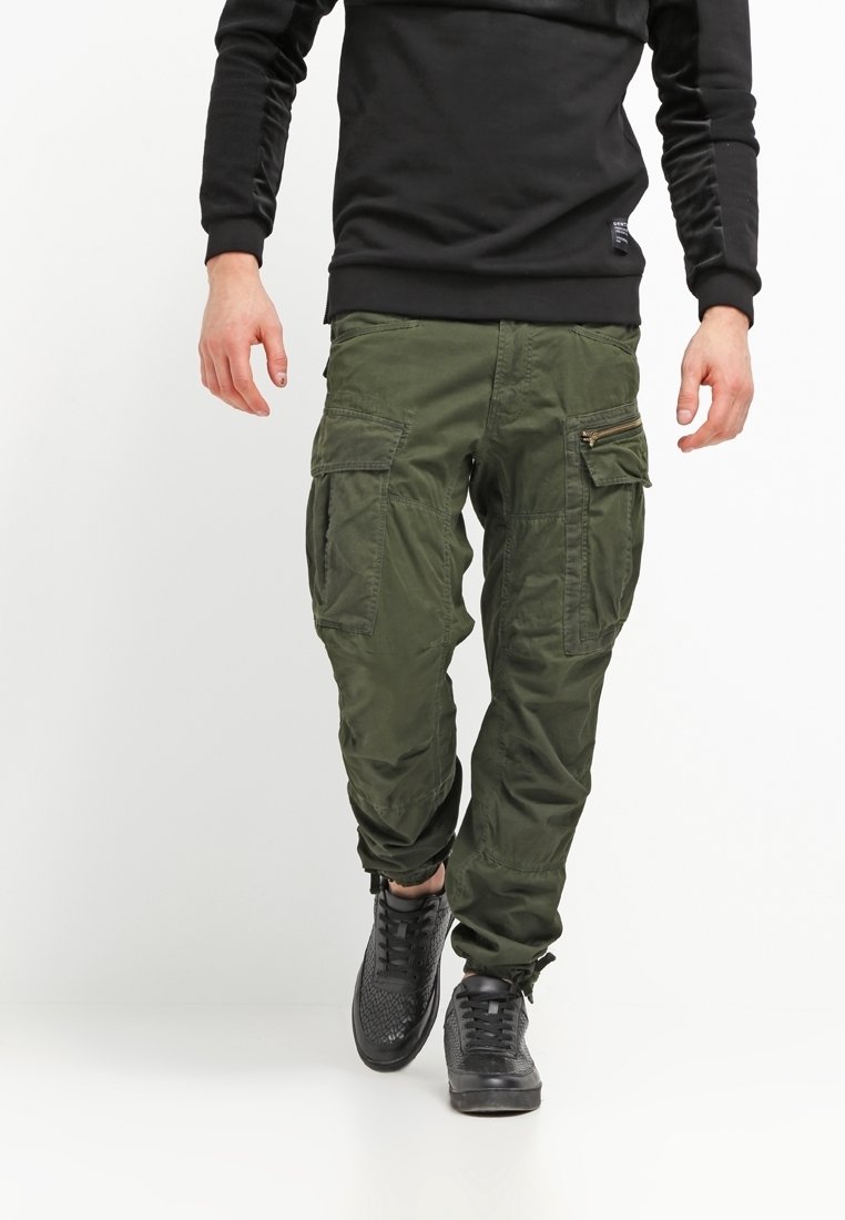 Gröna cargobyxor med flera fickor och dragkedjor, tillsammans med en svart långärmad tröja och svarta texturerade sneakers.