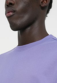 Close-up van het onderste deel van iemands gezicht en nek, met een lichtpaarse shirt met ronde hals, tegen een effen achtergrond.