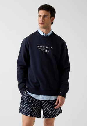 Mężczyzna noszący granatową bluzę z napisem „North Sails GUESS” na jasnoniebieskiej koszuli, w połączeniu z wzorzystymi granatowymi szortami.