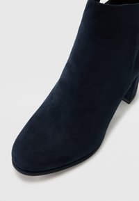 Tamnoplave čizme do članaka s visokom potpeticom, glatke zamršene teksture, zaobljenog vrha i čvrstog blok-peta. Nema vidljivih dijelova od metala ili ukrasa.