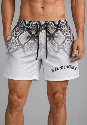 Uomo che indossa pantaloncini da bagno bianchi e grigi con motivo a serpente, coulisse nere e scritta "RICHMOND" sulla gamba sinistra.