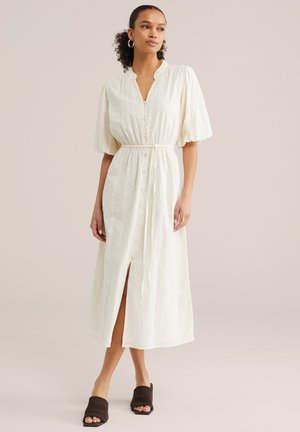 Donna che indossa un abito midi bianco con bottoni, maniche a sbuffo e cintura annodata in vita, abbinato a sandali mule neri con punta aperta.