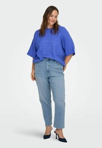 Donna sorridente con gli occhi chiusi che indossa un maglione azzurro a maniche corte largo, jeans azzurri chiari e sabot neri con tacco su sfondo bianco.